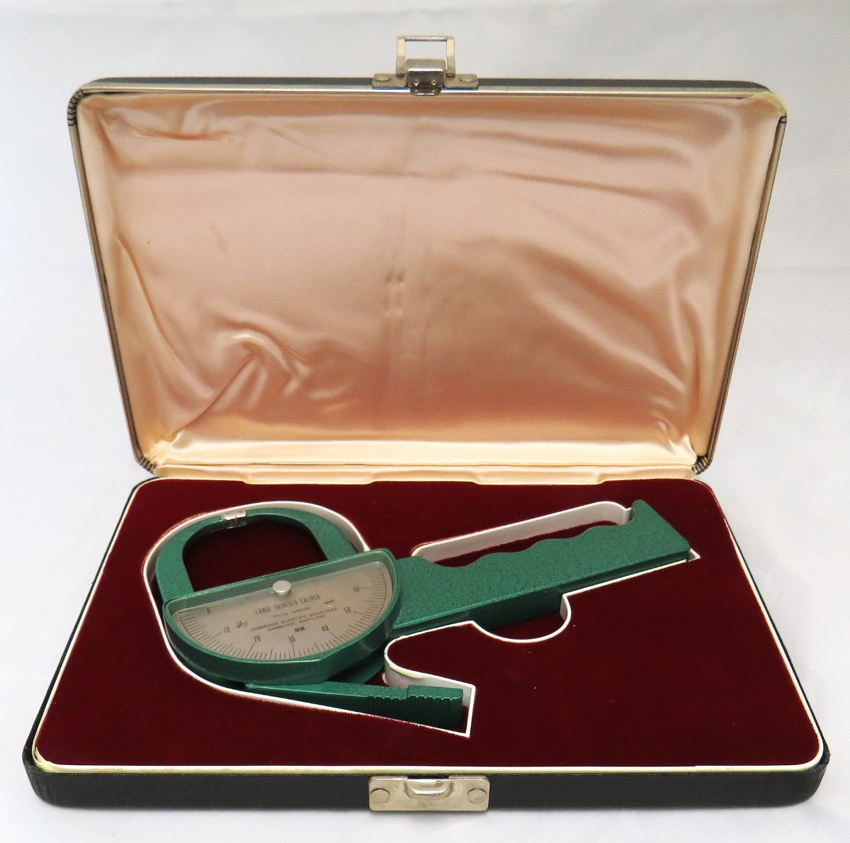 Green metal skinfold caliper in case lined with red velvet. The caliper has a dial on it to measure how far open it is.