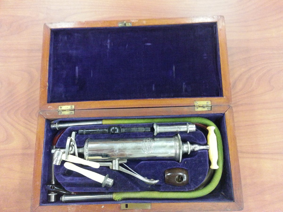 Wood case with blue velvet interior. Tools inside include a metal syringe with an ivory handle on the plunger, and a green rubber tube.