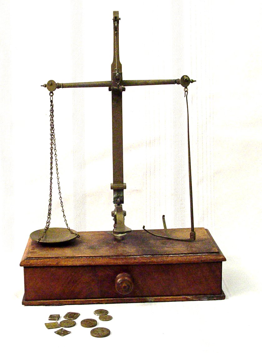 Apothecary scale made of wood base with drawer in it and an upright metal scale with round plates hanging on each side. In front are a bunch of tiny weights, both round and square shaped.