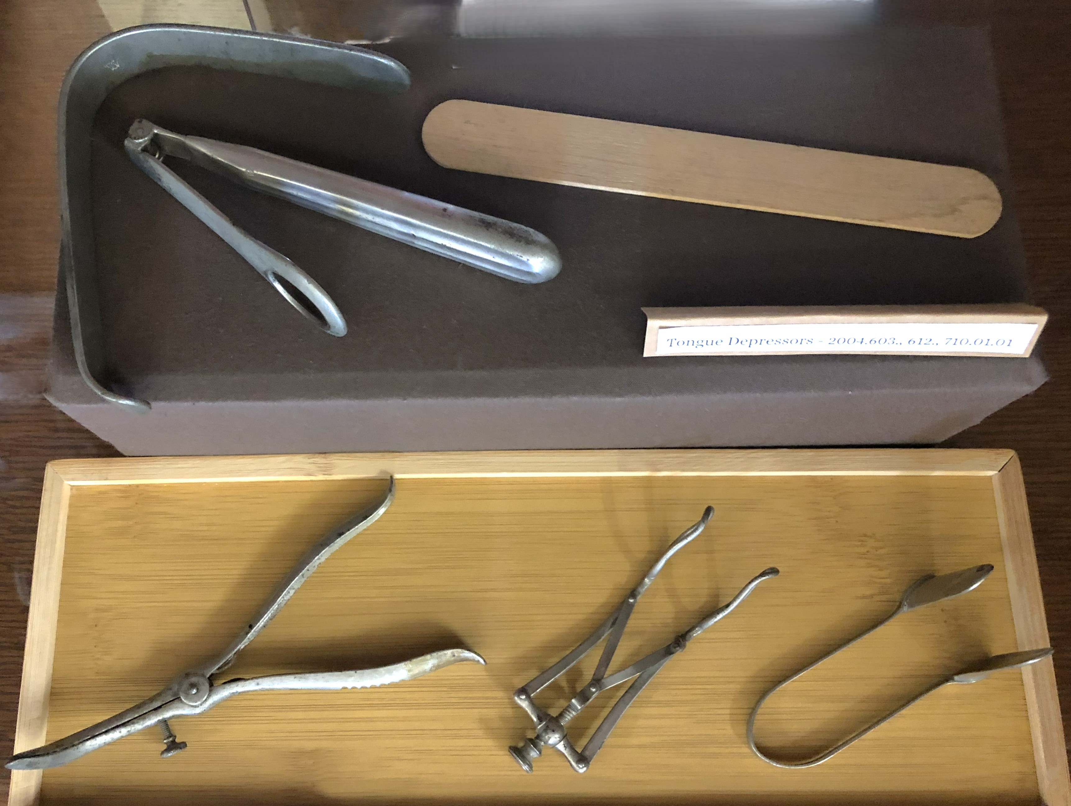 Three nasal specula, one a simple U shape, one with a screw and a spring, and one in the style of pliers; and three tongue depressors, one a wooden popsicle stick, one foldable with a fenestrated end, and one a bent piece of metal.