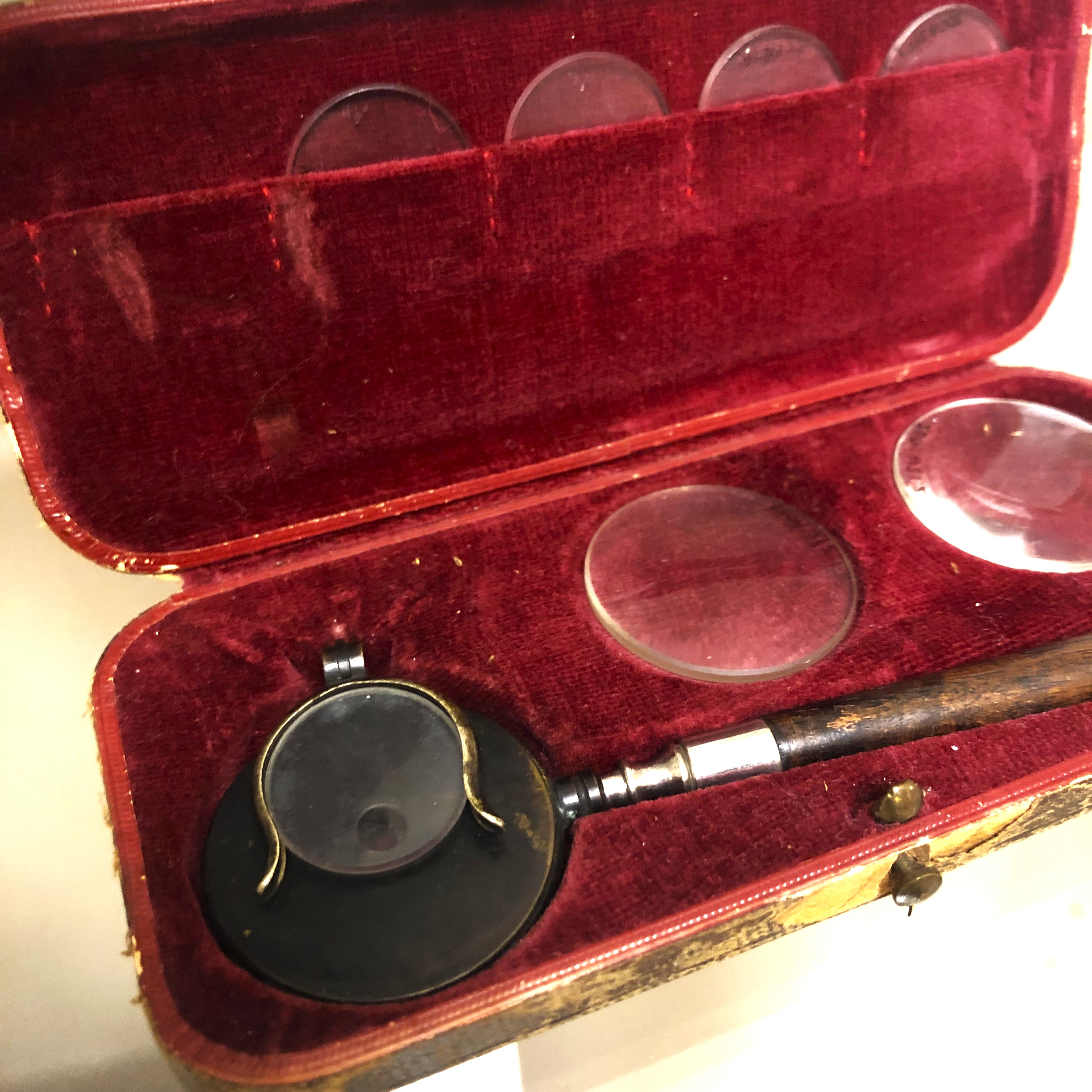 Red velvet case with round black instrument inside with long thin handle; extra lenses are beside.